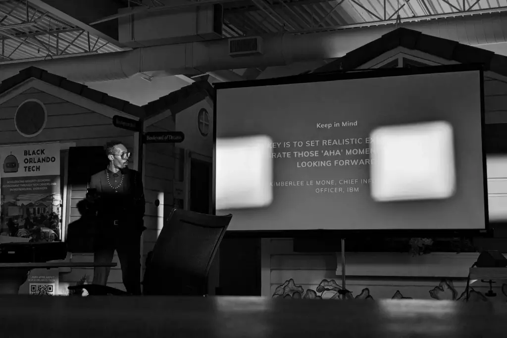 David standing small beside a projector screen in a dim, beamed room
